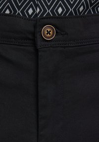 Tmavé kalhoty v pase s hnědým knoflíkem a černobílou geometrickou vzorovanou látkou viditelnou nad páskem.