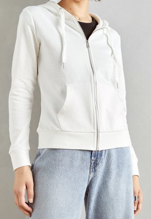 Person wearing a white zip-up hoodie with front pockets and blue jeans, standing against a plain gray background.