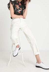Pantaloni bianchi a vita alta con risvolti, abbinati a una blusa floreale scura con maniche arricciate e scarpe bianche con accenti neri.