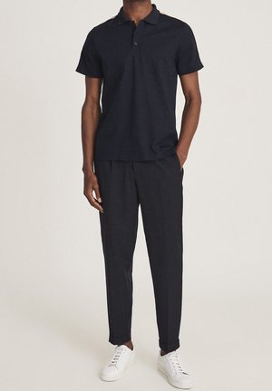 Homme portant un polo noir à manches courtes, un pantalon noir avec des revers, et des baskets blanches, debout, une main dans la poche.