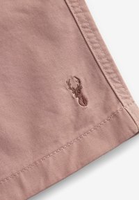 Coin de tissu rose poussiéreux avec logo tête de cerf brodé et couture double sur fond blanc.