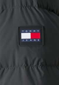 Zwarte pufferjack-stof met een gestikte textuur. Bevat een rechthoekig label met "TOMMY JEANS" in witte letters en een rood-wit logo.