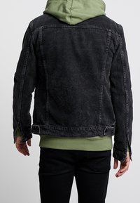 Chaqueta de denim negro con textura desgastada, que incluye una sudadera verde con capucha debajo. Presenta costuras visibles y aberturas laterales.