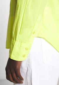 Main à la peau foncée à côté de la poche d'un pantalon blanc, vêtue d'une chemise jaune vif à manches longues avec un poignet boutonné.