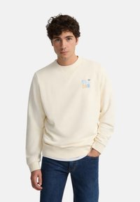 Joven con cabello rizado, vistiendo un sudadero color crema con un pequeño logo colorido en el pecho y jeans azules, con una mano en el bolsillo.