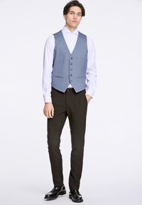 Blåmönstrad väst med knappar, buren över en vit skjorta, i kombination med svarta byxor och glänsande svarta skor. Enkel, skräddarsydd design.