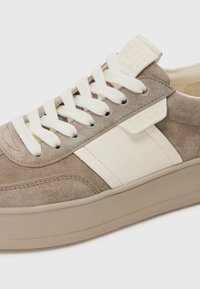 Sømsneakers i lys brun med en kremfarget lærstripe, som har hvite snørebånd, en teksturert hælklaff, og en flat, beige gummisåle.