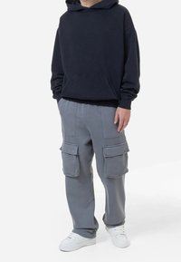 Person som bär en svart hoodie, grå cargobyxor med stora fickor och vita sneakers, stående mot en vit bakgrund.