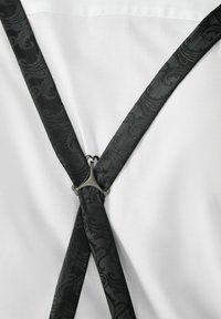 Mustad mustriga traksid dekoratiivse mustriga, millel on hõbedane metallklamber, risti valge tekstuuriga kangast särke peal.