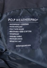 Laadukkaasta tummansinisestä kankaasta valmistettu etiketti, jossa on valkoinen teksti. Tiedoissa mainitaan vedenpitävyys, hengittävyys, tuulenpitävyys, kestävyys, kierrätetty vuori ja 360° heijastavuus.