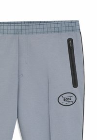 Grå atletiska byxor med en texturerad yta, elastisk midja och en ficka på sidan med dragkedja som har svarta accenter och en logotyp. 