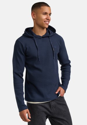 Mörkblå långärmad hoodie i ribbat tyg, med en framficka, justerbar dragsko på huvan och en subtil logotyp. Stylad med mörka jeans.