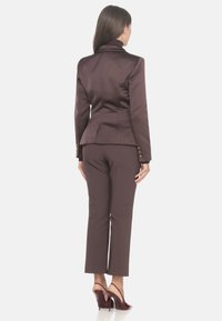 Blazer marrone aderente con rever a punta, vita sagomata e dettagli in oro, abbinato a pantaloni marroni coordinati e tacchi bordeaux.