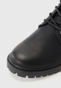 Bota de cuero negro con punta redondeada, superficie texturada y suela de goma gruesa con un patrón de relieve. Los cordones son oscuros con ojales metálicos.