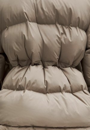 Detailný záber na materiál v taupe farbe so prešitými a vypolchovanými časťami, pravdepodobne súčasť vystuženej zimnej bundy alebo kabátu.