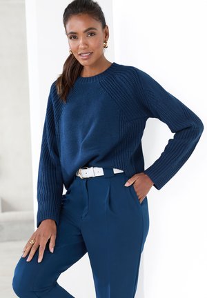 Marineblauer Strickpullover mit Rippendetails und Rundhalsausschnitt, kombiniert mit hochgeschnittenen marineblauen Hosen und einem weißen Gürtel. Keine Muster oder Akzente.