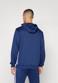 Sweat à capuche bleu marine avec des poignets et une bordure côtelés, présentant des rayures blanches sur les côtés et une texture lisse. Vue de dos, montrant les détails de la capuche.