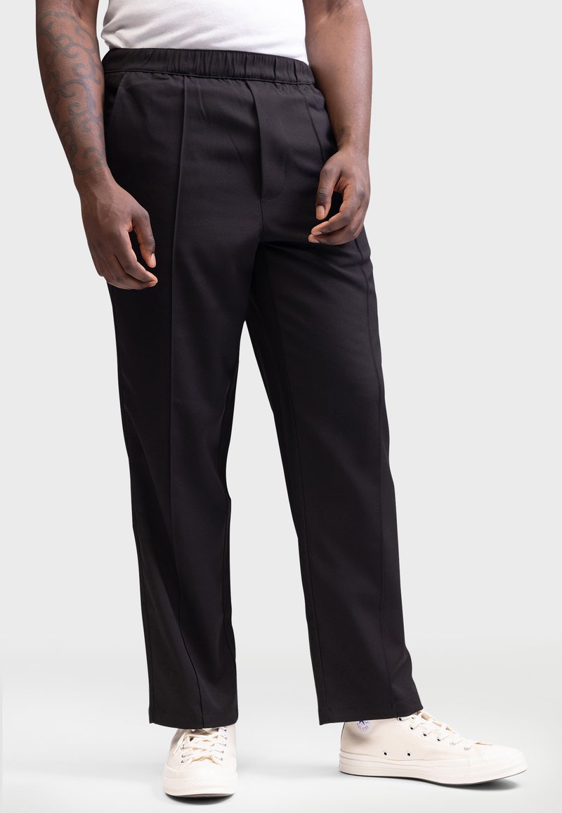Pantalons noirs ajustés avec une taille élastique, présentant une texture lisse et une coupe droite, associés à des baskets blanches.