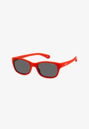 Lunettes de soleil en plastique rouge avec des montures rectangulaires, verres gris et texture lisse. Présentent un logo sur la branche.