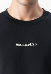Svart bomullssweatshirt med rund hals og hvit brodert "marimekko"-logo på brystet. Teksturert stoff, enkel design.
