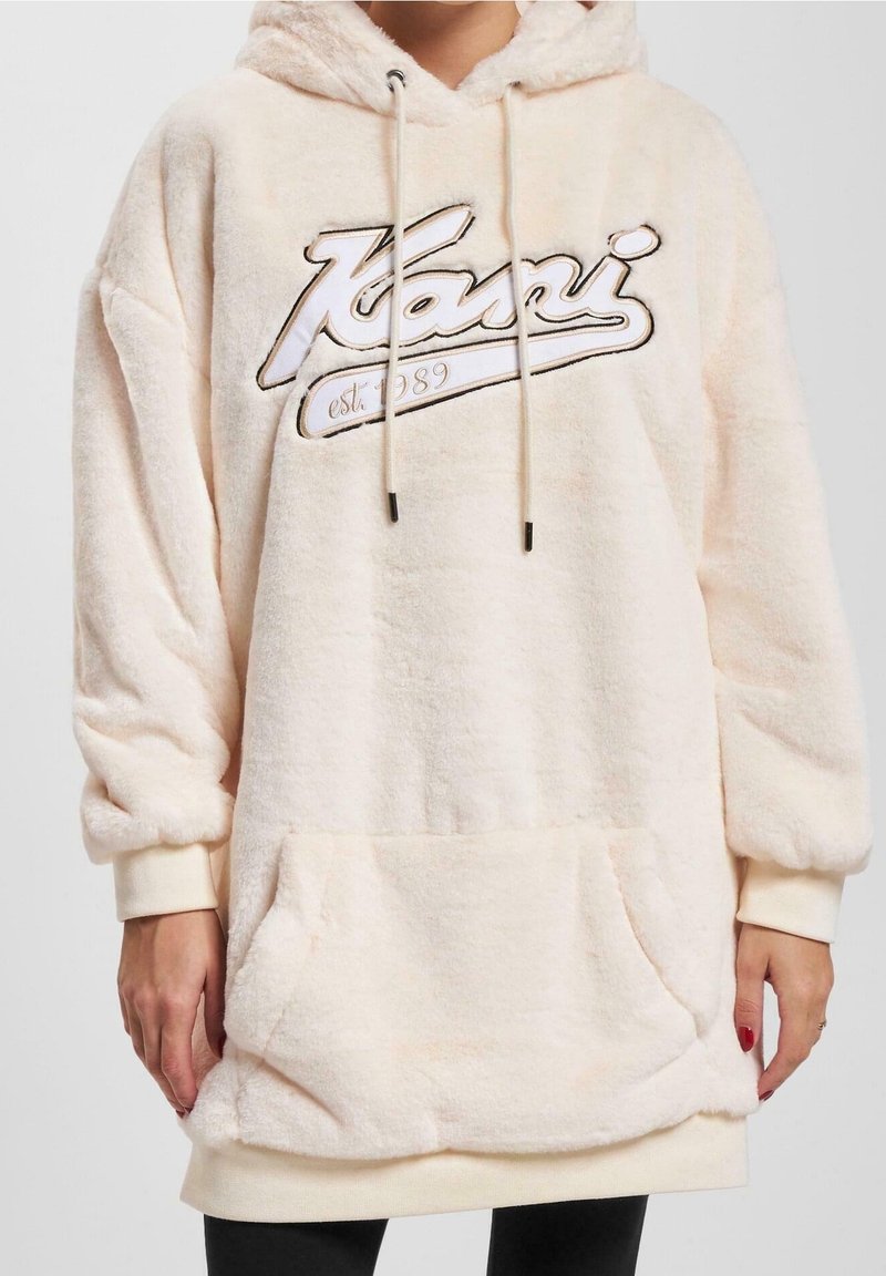 Personne portant un sweat à capuche oversize beige clair et moelleux, avec une poche avant et des cordons, arborant le texte « Kani est. 1989 » sur la poitrine.