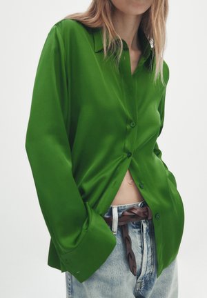 Femme portant une chemise ample en satin vert vif à boutons, partiellement rentrée dans un jean bleu clair avec une ceinture marron tressée.