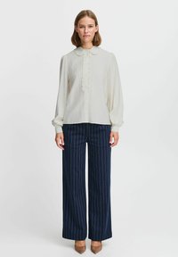 Weiße Bluse mit raffiniertem Kragen und Rüsche, kombiniert mit marineblauen, gestreiften weiten Hosen und beigen Absatzschuhen. Glatte Stofftextur.