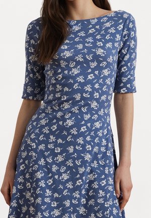 Blå kjole med hvidt blomsterprint, figursyet talje og ærmer til albuen båret af en person med langt brunt hår.