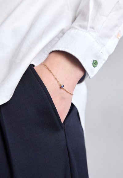 Gold bracelet featuring a small blue gemstone, worn on a wrist beside a white shirt with green and orange buttons and navy pants.