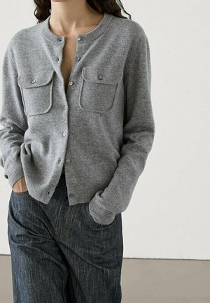 Femme portant un cardigan gris clair boutonné avec deux poches à rabat à l'avant et un pantalon ample gris foncé taille haute, les mains dans les poches.