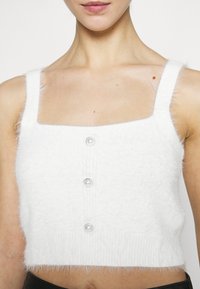 Top corto bianco realizzato in materiale morbido e peloso. Presenta spalline larghe, scollo quadrato e tre bottoni simili a perle lungo la parte anteriore.