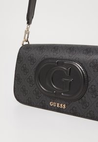 Schwarze Handtasche aus strukturiertem Material mit einem auffälligen geprägten Logo, metallischen Beschlägen und einem verstellbaren Tragegurt.