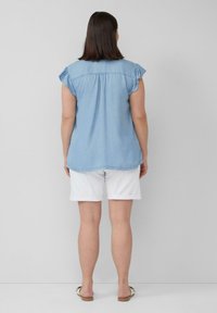 Blaues Denim-Oberteil mit kurzen Ärmeln und Rüsche an den Schultern, mit abgerundetem Saum. Kombiniert mit weißen hochgekrempelten Shorts und flachen Sandalen.