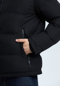 Czarna kurtka puchowa o gładkiej teksturze, z zamykaną kieszenią po lewej stronie i dopasowanym mankietem na rękawie.