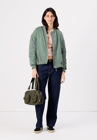 Chaqueta bomber verde claro con puños acanalados, top corto rosa, jeans oscuros de cintura alta y un bolso verde oliva con dobles bolsillos.