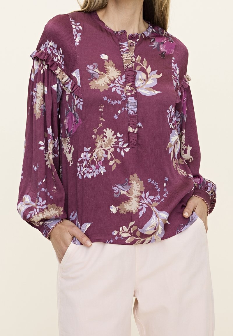 Femme portant un chemisier floral prune à manches longues avec des détails à volants, associé à un pantalon beige clair, mains dans les poches.