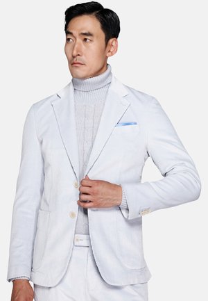 Man wearing a light blue corduroy suit with a blue pocket square and a gray cable-knit turtleneck sweater, looking to the side.