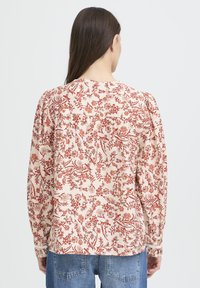 Femme aux longs cheveux bruns portant un haut crémeux avec des motifs floraux rouges et un pantalon en jean bleu, se tenant face à un fond blanc.