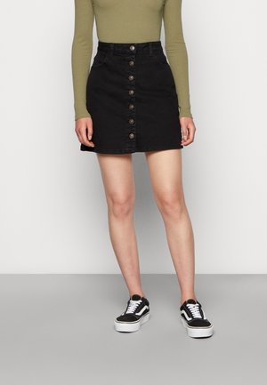 Femme portant une jupe en denim noire boutonnée à l'avant, un haut à manches longues olive et des baskets noires et blanches, debout devant un fond uni.