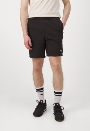 Zwarte katoenen shorts met een elastische tailleband, voorzien van zijzakken en een wit logo. Draagt met zwarte schoenen en gestreepte witte sokken.