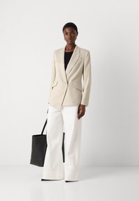 Beige bleiser med strukturert design, to frontlommer, båret over en svart topp. Et par hvite vide bukser og en svart tote bag.