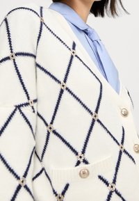 Cárdigan de punto en color crema con un patrón de diamantes en azul marino y botones dorados con flor; presenta un cuello en V y bolsillos con bordes acanalados.