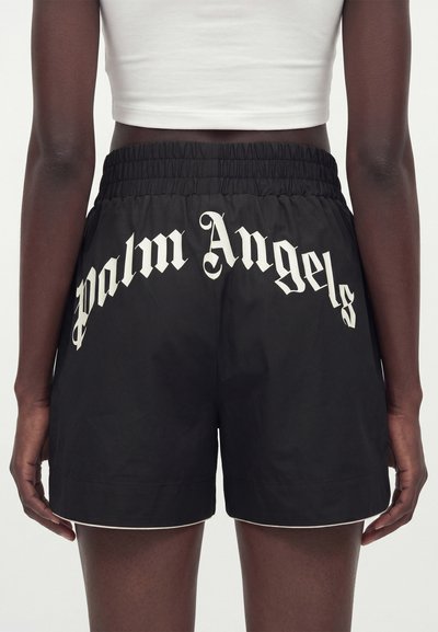Άτομο που φοράει μαύρα σορτς με λευκό κείμενο "Palm Angels" κυρτωμένο στην πίσω μέση, σε συνδυασμό με λευκό κοντό τοπ.