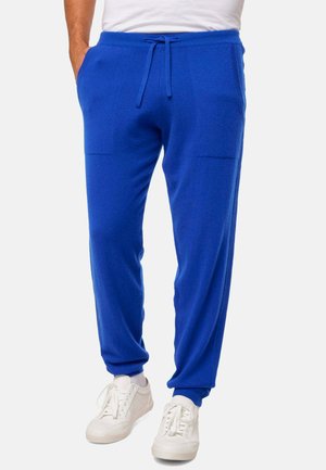 Homme portant un pantalon de jogging bleu vif avec cordon de serrage et des baskets blanches, se tenant avec les mains dans les poches.