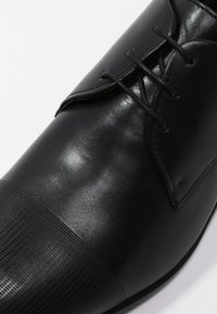 Chaussure en cuir noir avec une tige lisse, un bout arrondi et des lacets. Le bout présente un motif texturé pour plus de détails.