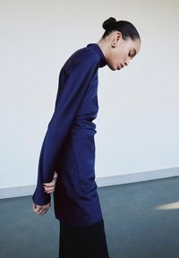 Top en satin bleu marine à manches longues, col montant et fente latérale. Texture lisse et coupe ajustée mise en valeur par une jupe noire contrastante.