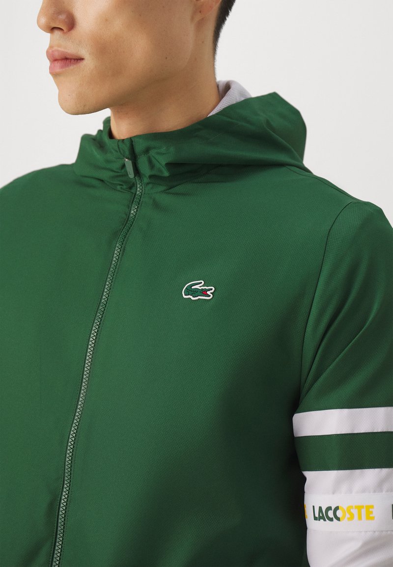 Grüne Zip-Hoodie aus strukturiertem Material. Verfügt über eine Kapuze, weiße Streifenakzente an den Ärmeln und ein kleines Lacoste-Logo auf der Brust.
