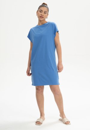 Vrouw staand, gekleed in een blauwe jurk met korte mouwen tot op de knie en beige instappers, tegen een effen witte achtergrond.