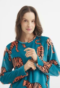 Camiseta de forro polar azul con mangas largas que presenta patrones de tigre naranja. Textura suave, escote redondo y un ajuste relajado.