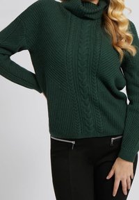 Pull à col roulé vert foncé avec une texture côtelée et un motif en torsades. Pantalon noir avec poches zippées.